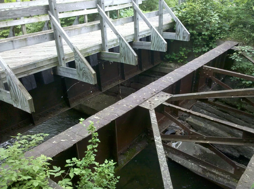 Remains of an abandoned metal bridge with rusty girders and broken planks above a stream, beside a wooden footbridge.