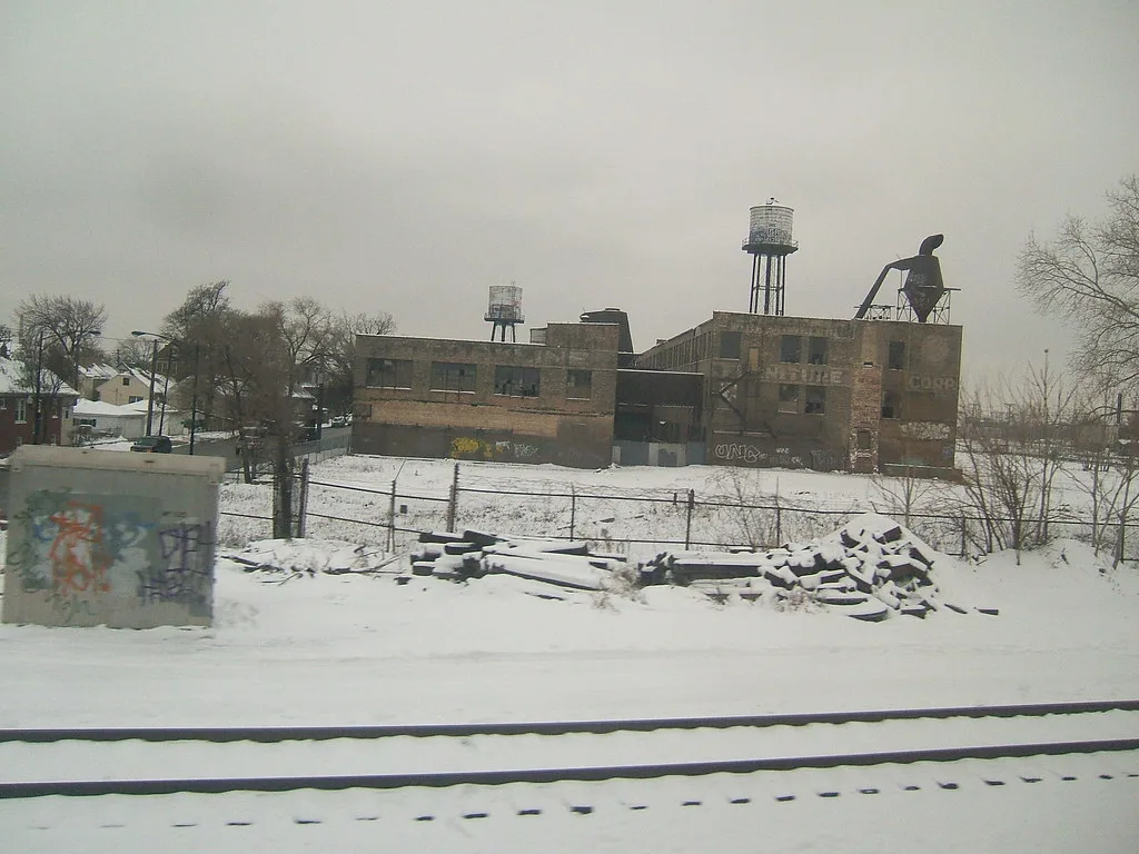 Abandoned brick factory with broken windows and rooftop tanks behind snow-covered railway tracks.