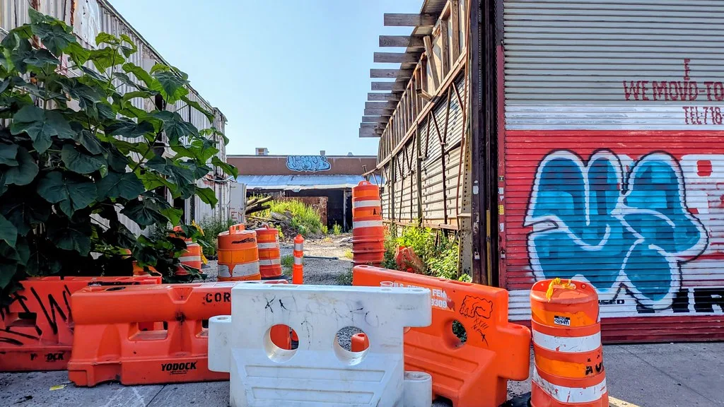 Passage industriel abandonné entre bâtiments dégradés, avec plantes, charpente découverte et barrières orange devant une porte graffitée.