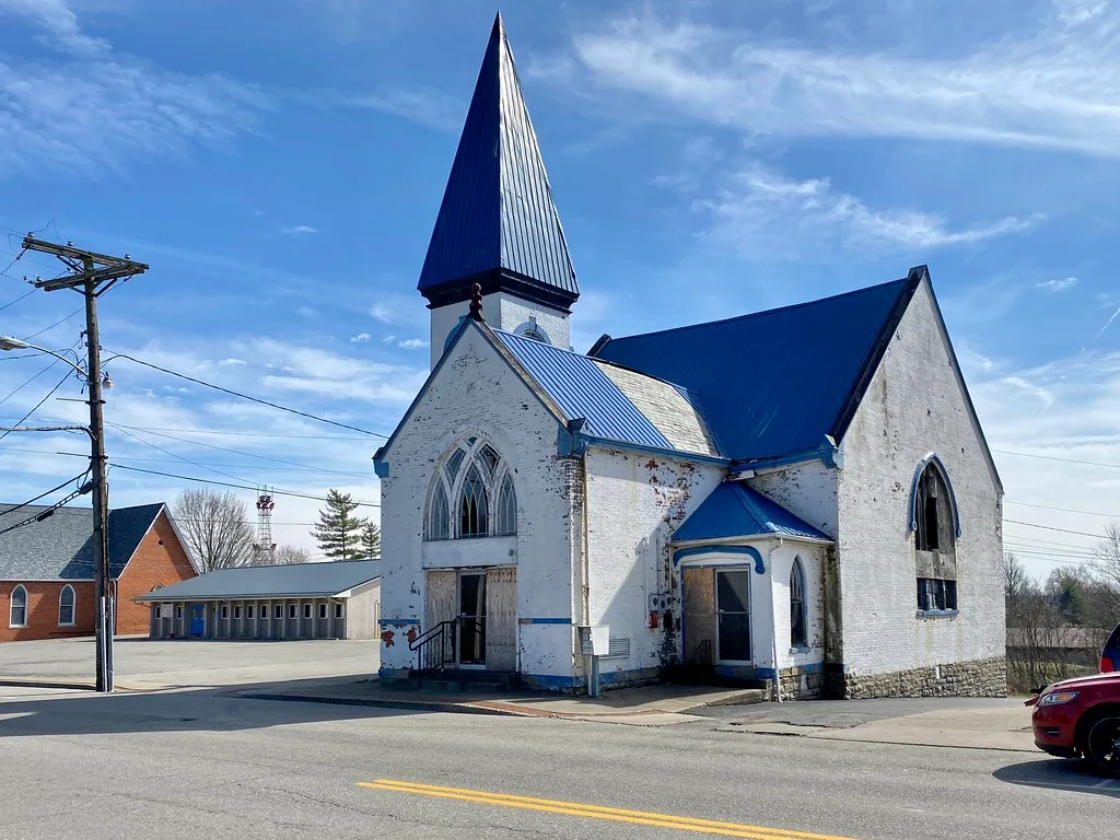 Old white church with a blue roof and partly boarded openings.