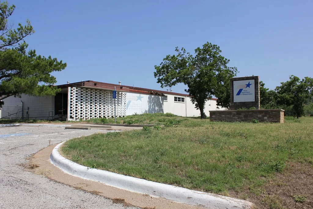 Low former administrative building beside an empty grassy parking area.