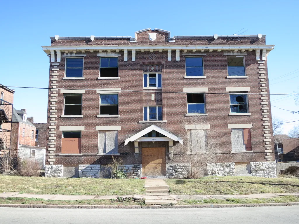 Abandoned three-storey brick apartment building with shattered panes and a wood-boarded entrance.