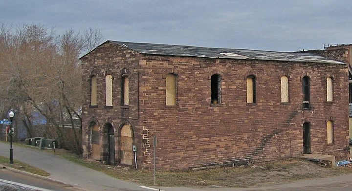 Long brown stone building with boarded or glassless windows and a damaged roof.