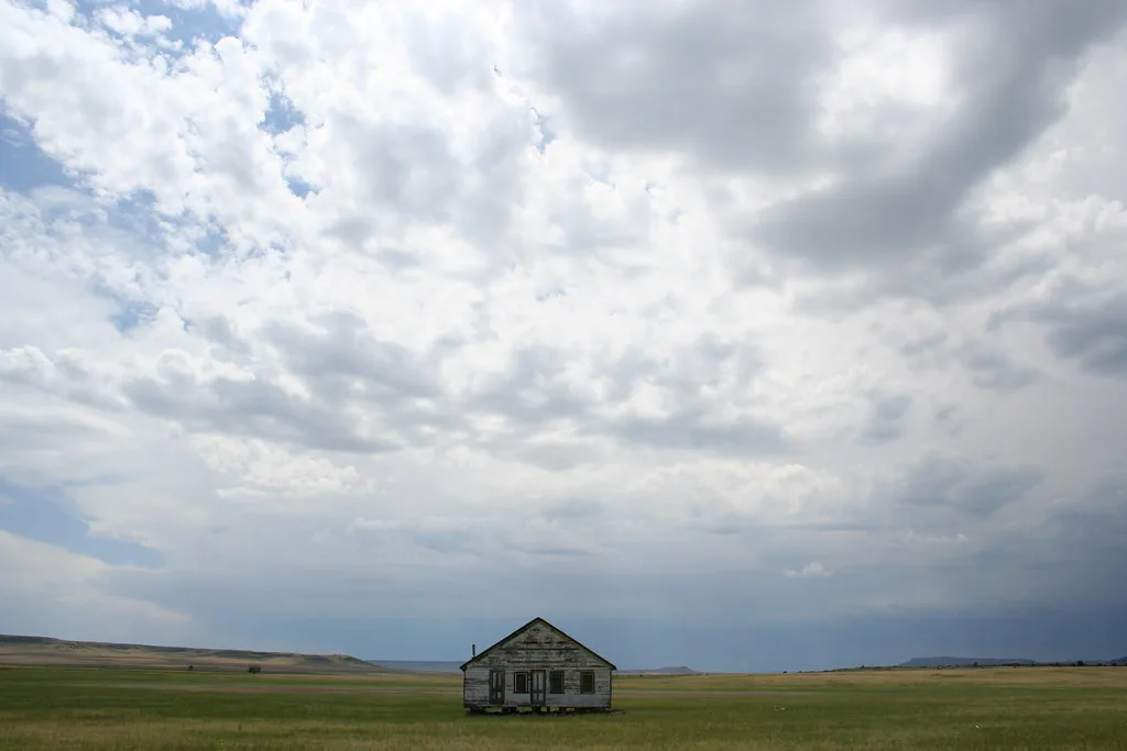 Old wooden house standing alone in a broad meadow beneath a cloudy sky.
