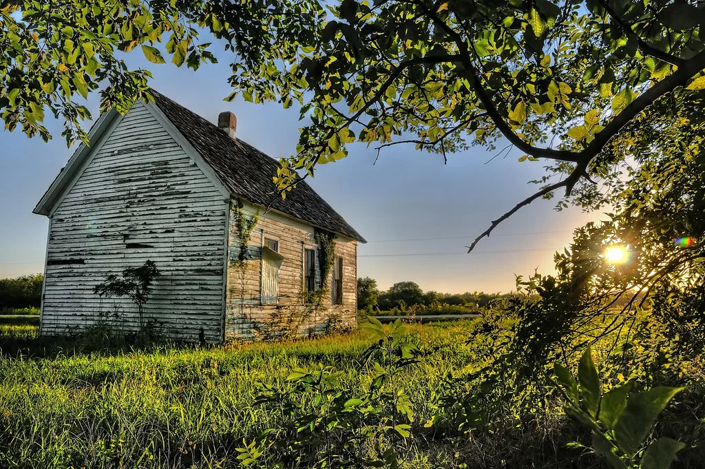 White wooden house with boarded windows and peeling paint amid grass at sunset.