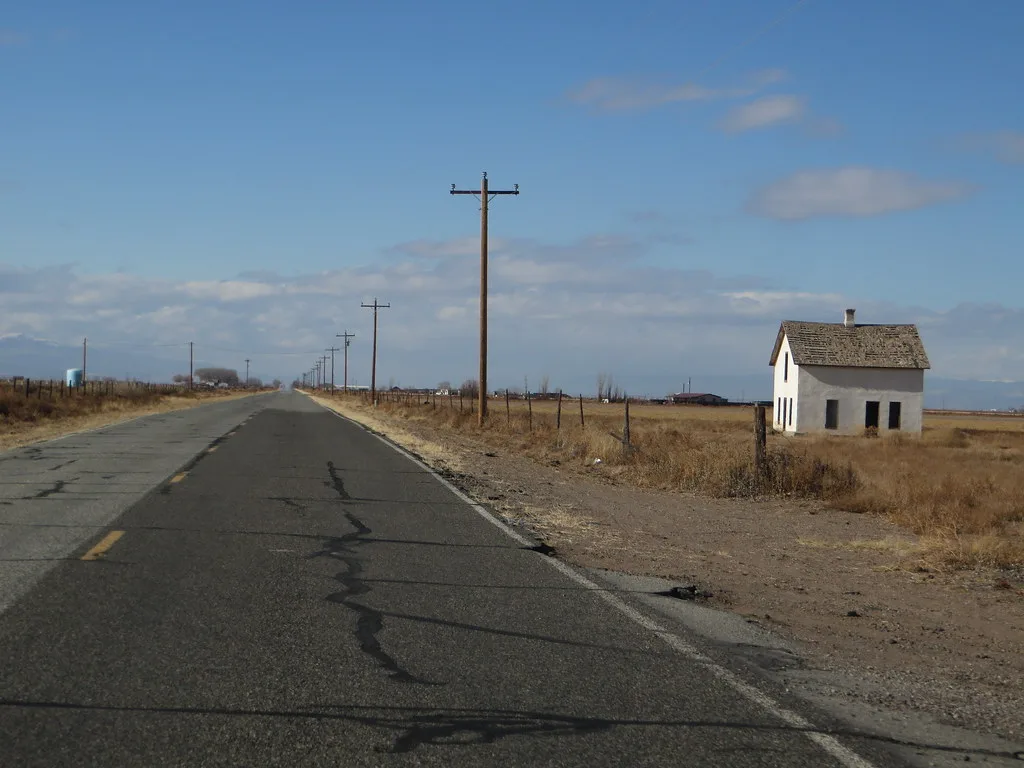 Petite maison abandonnée blanche aux fenêtres vides près d’une longue route droite dans une plaine sèche.