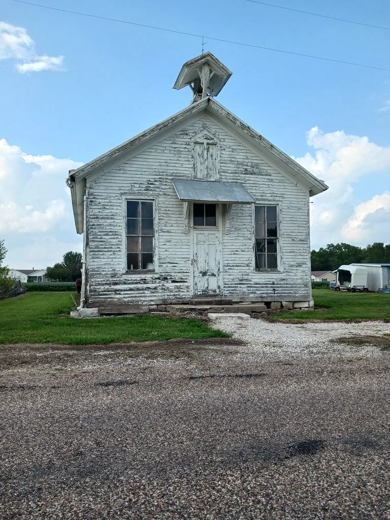 Small abandoned chapel with peeling white timber, broken windows and a wooden bell turret above the entrance.