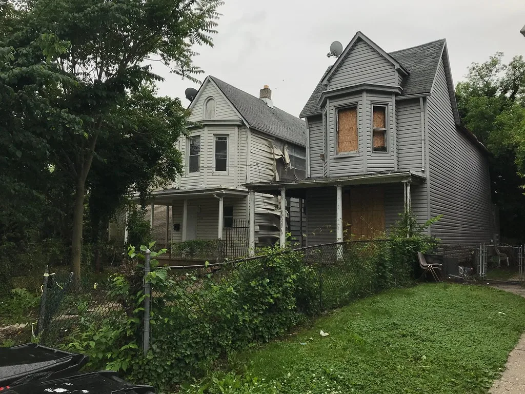 Two grey abandoned houses with empty or boarded windows and warped cladding behind an overgrown fence.