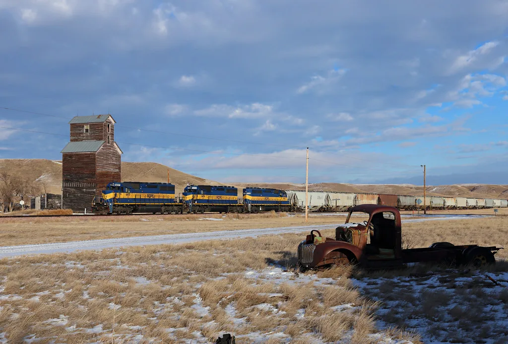 Old wooden grain elevator beside a railway, with a train and a truck wreck.