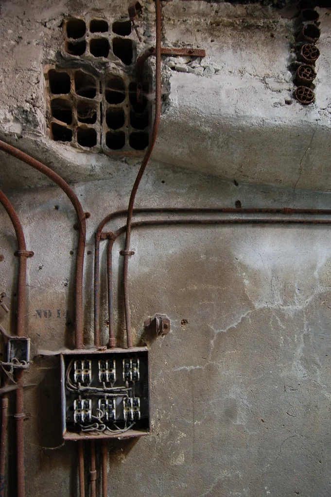 Ancien tableau électrique ouvert et conduits rouillés sur un mur effrité.