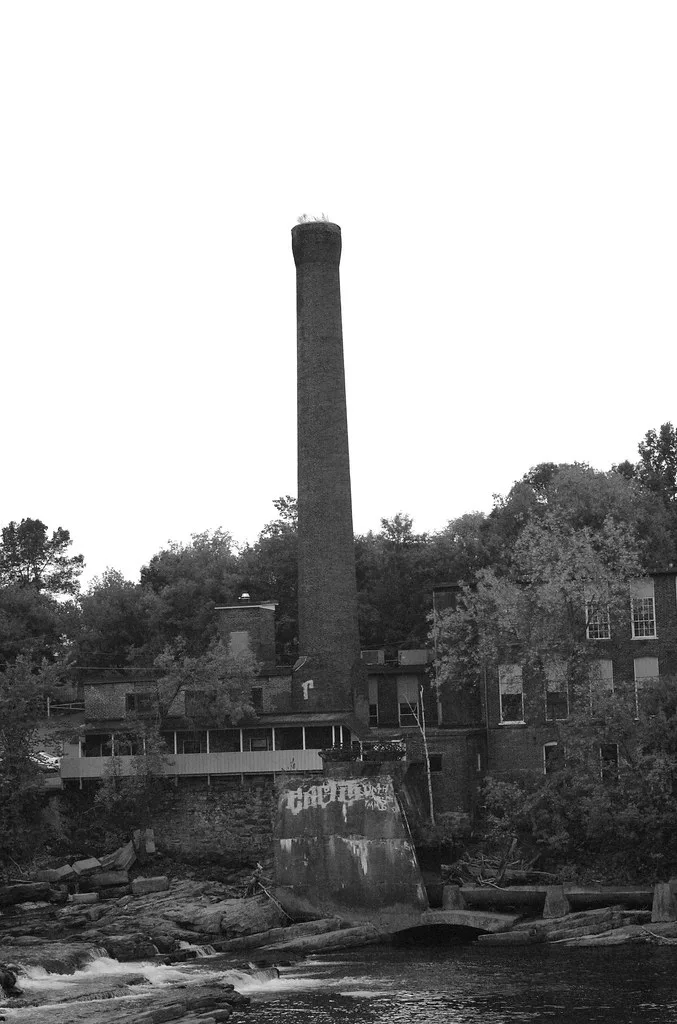 Abandoned factory with a tall brick chimney beside a rocky river.