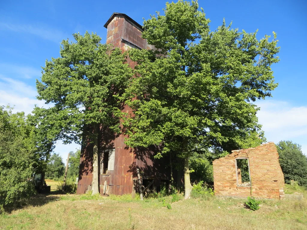 Rusty industrial tower between trees beside a roofless remnant of a brick wall.