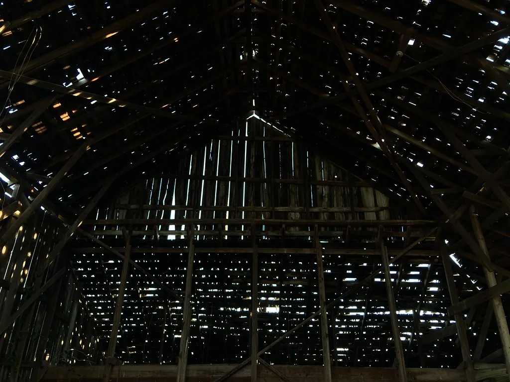 Interior framing of a dark abandoned barn, with light passing through numerous holes in its roof and wooden cladding.