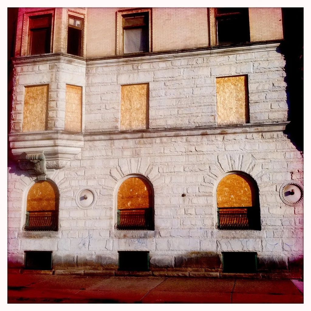 Façade abandonnée de pierre claire aux fenêtres cintrées condamnées et baies supérieures cassées.