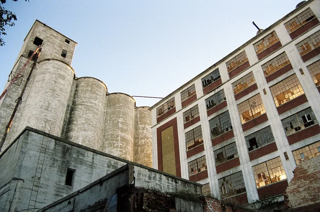 Concrete silos before a factory with many broken windows and eroded walls.