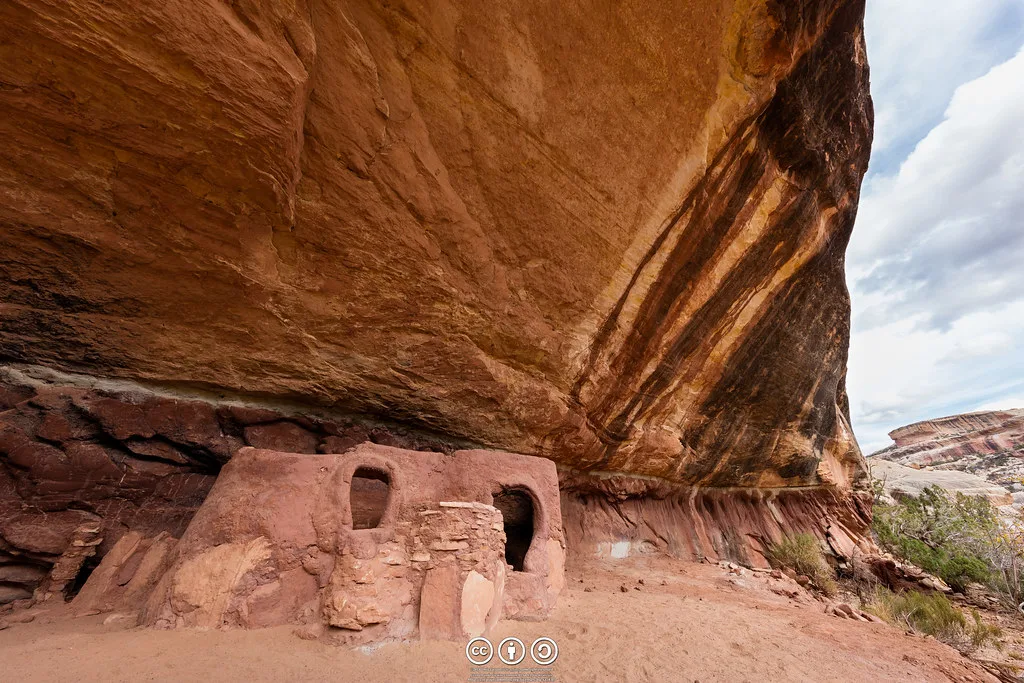 Small ruined red-earth rooms beneath a vast sandstone overhang.