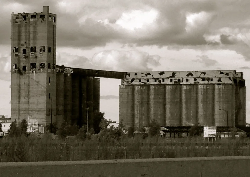 Dilapidated old concrete silos beneath a cloudy sky, in sepia.