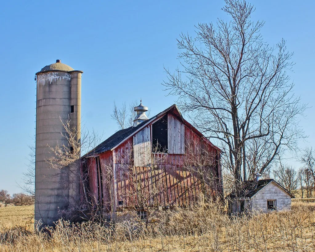 Abandoned red barn with separated boards beside a cylindrical silo and a ruined annex in brush.