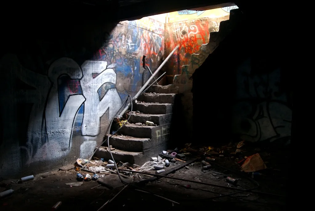 Damaged concrete staircase rising from a dark basement covered in graffiti and debris.