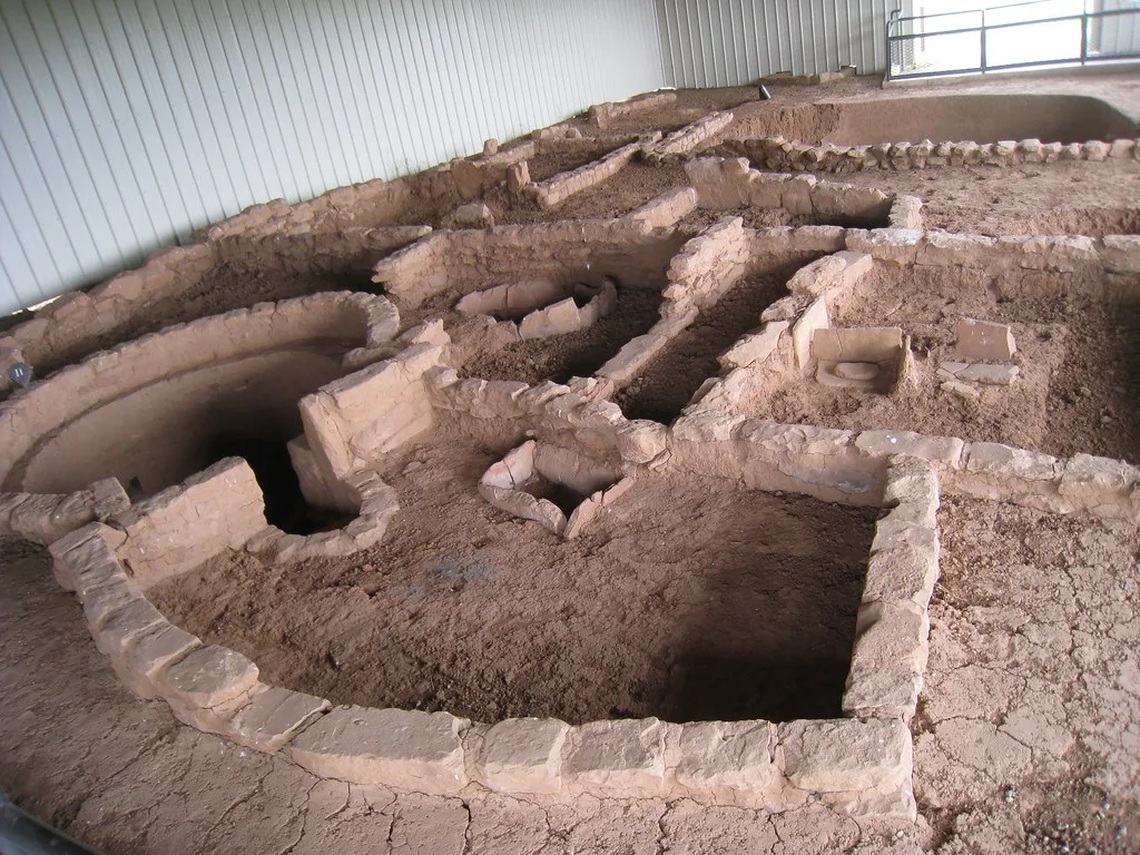 Vestiges de petites pièces en pierre et terre avec murs arasés et cavités circulaires sous un abri.