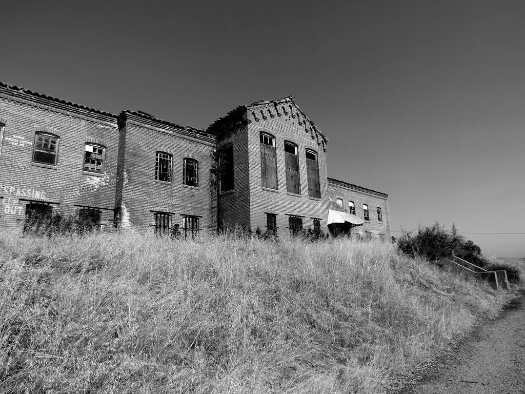 Closed old brick building surrounded by tall grass, in black and white.