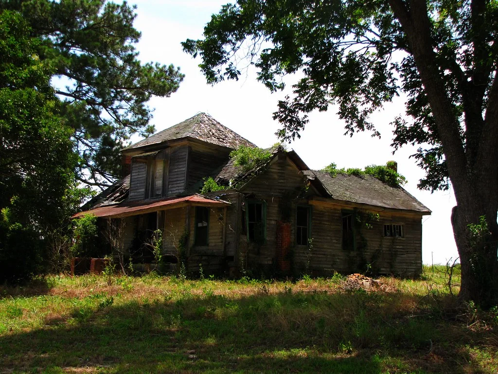 Large dark wooden house with empty windows and a sagging plant-covered roof.