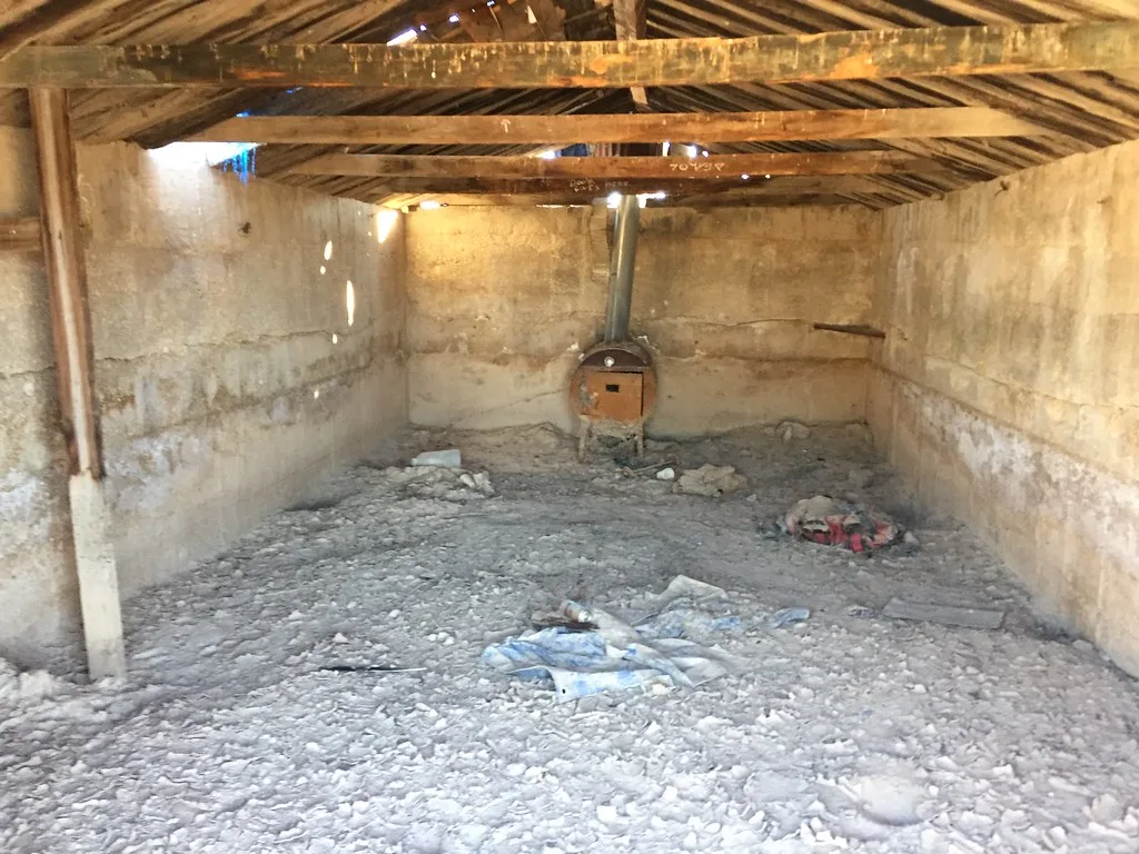 Dilapidated room beneath a perforated wooden roof with an old stove at the back.