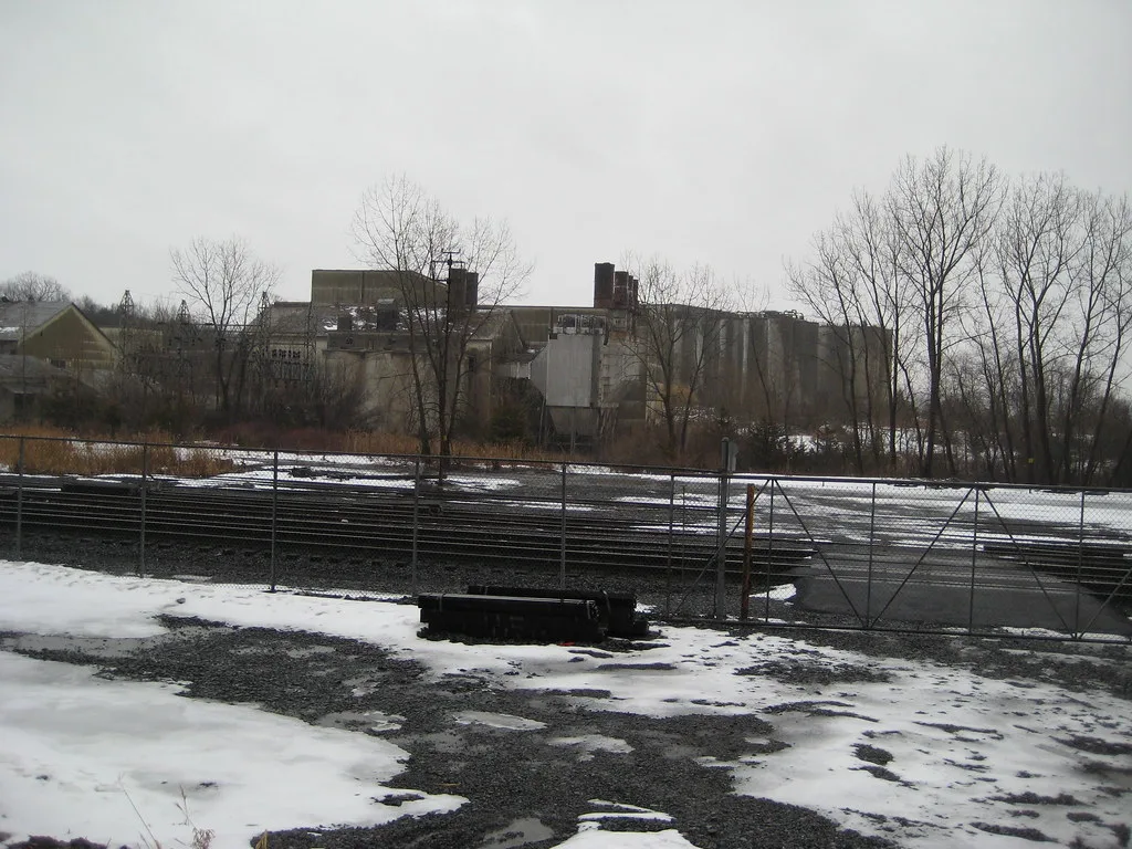 Abandoned former factory and silos behind a fence and snow-covered rails among bare trees.