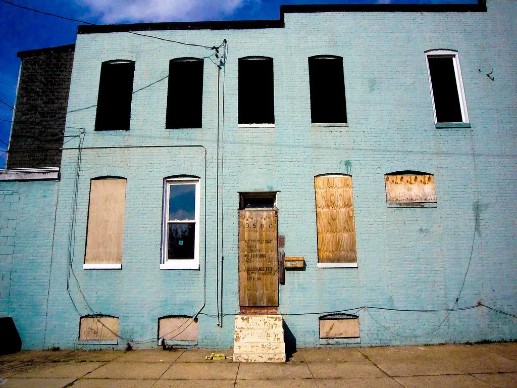 Pale blue facade of an abandoned building with empty windows and barricaded doors.