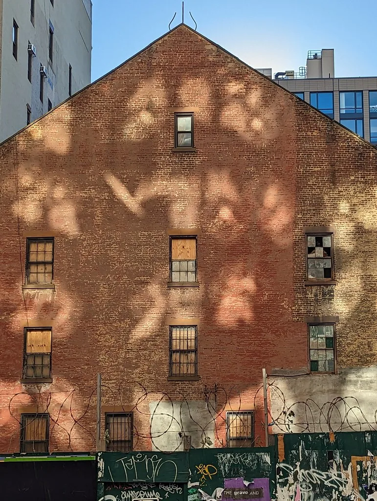 Tall brick gable with boarded or broken windows behind a graffiti-covered fence and barbed wire.
