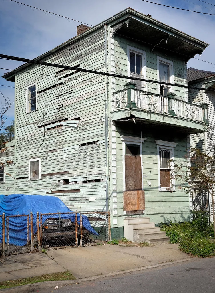 Abandoned pale green wooden house with a balcony, missing boards and broken or boarded windows.