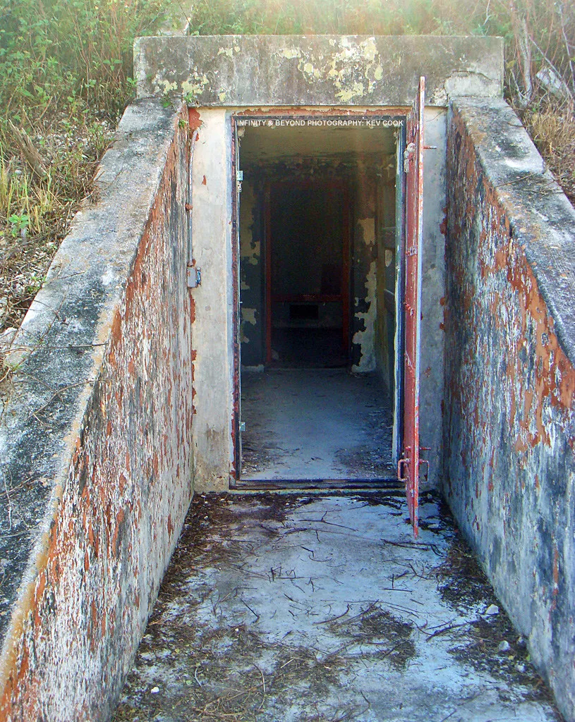 Sunken concrete entrance to an underground room, with an open metal door and peeling paint.