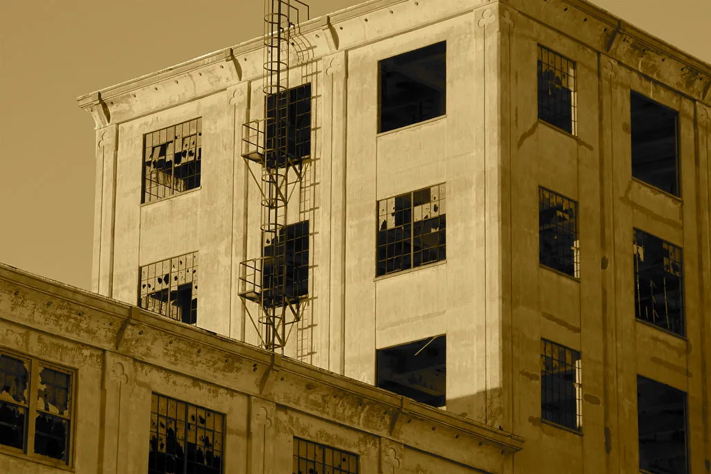 Old industrial building with broken windows and an exterior ladder, in sepia tones.