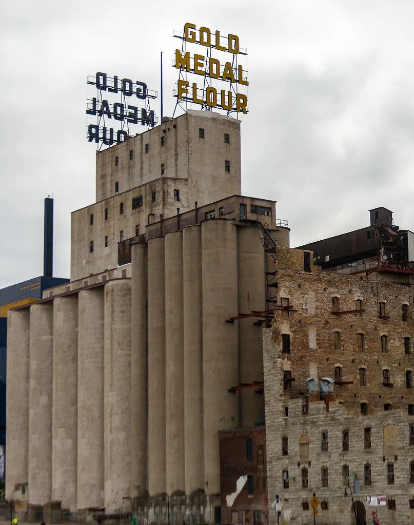 Concrete silos beneath an old flour sign, beside the broken walls of a roofless industrial building.