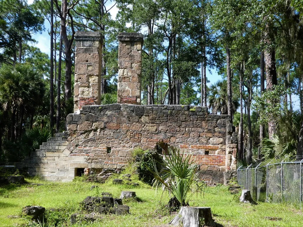 Two stone chimneys rising above ruined walls among pine trees.