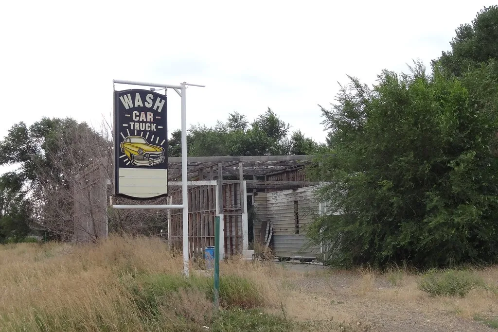 Abandoned former car wash with an exposed wooden frame behind its sign, amid tall grass.