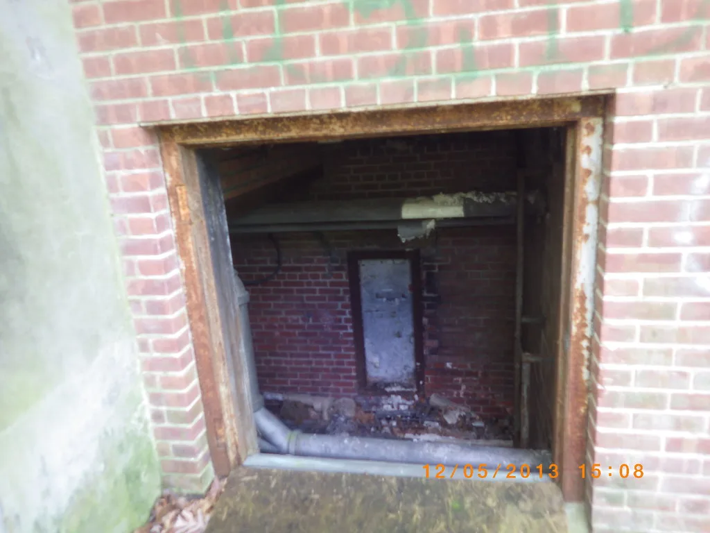 Ouverture rectangulaire rouillée donnant sur une pièce de briques abandonnée avec tuyau tombé et débris.