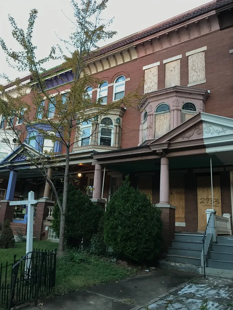 Row house with boarded doors and windows behind bushes.