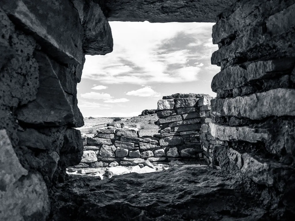 Collapsed walls of an abandoned ruin seen through an unglazed stone opening, in black and white.