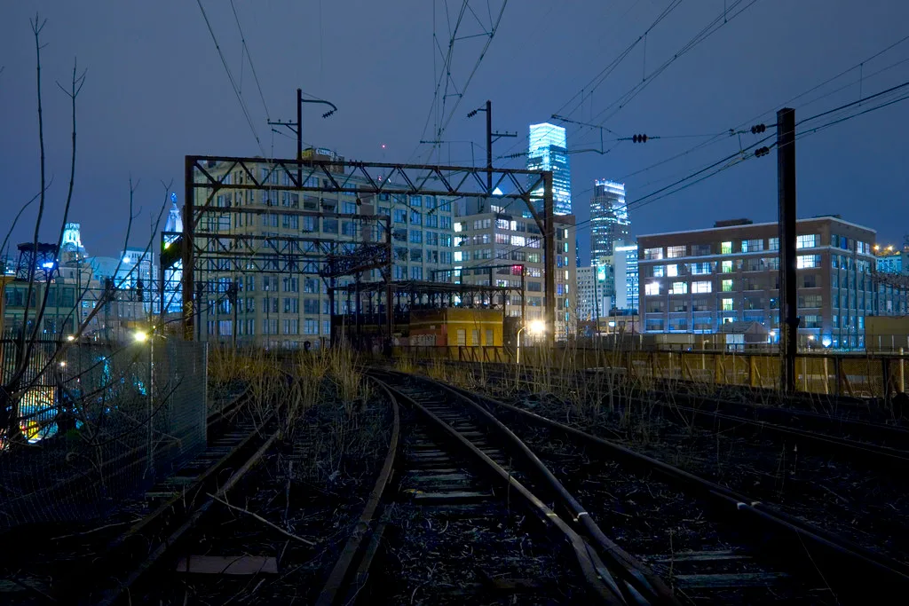 Voies ferroviaires délaissées envahies de jeunes arbres devant des immeubles éclairés de nuit.