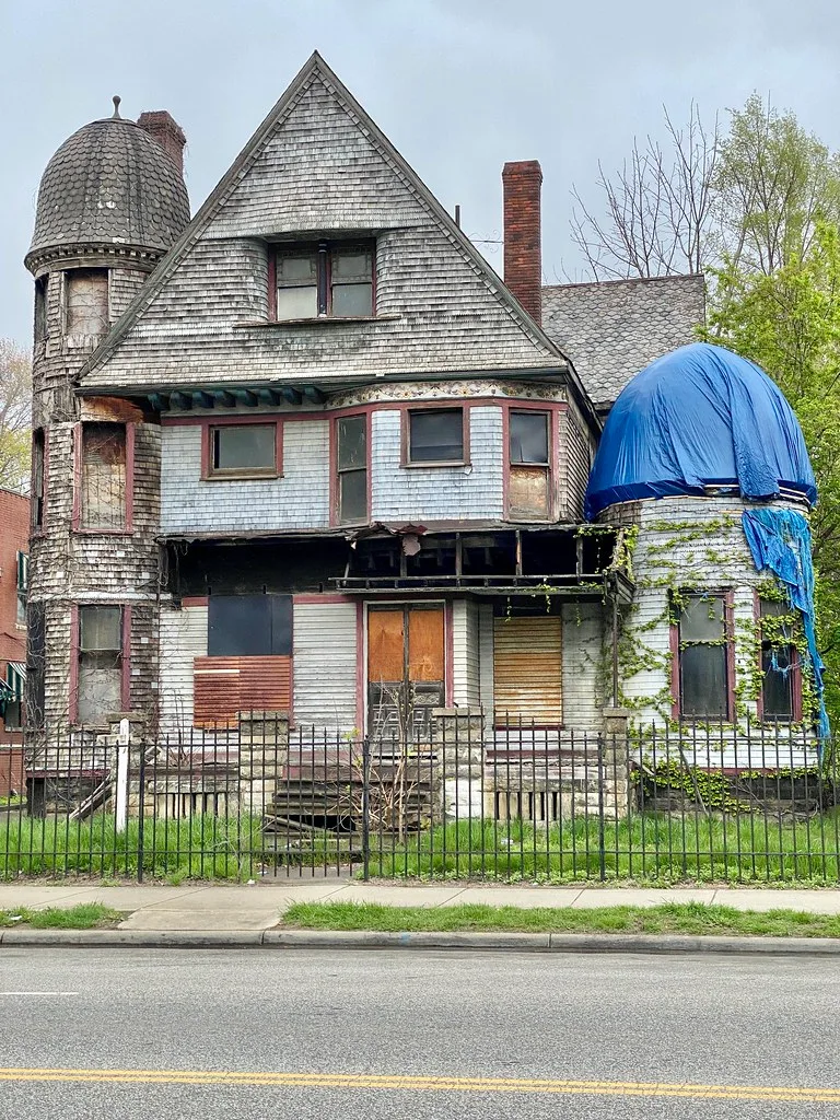 Maison abandonnée à tourelles et bardage gris, fenêtres condamnées et porche détruit, avec une tourelle sous bâche bleue.