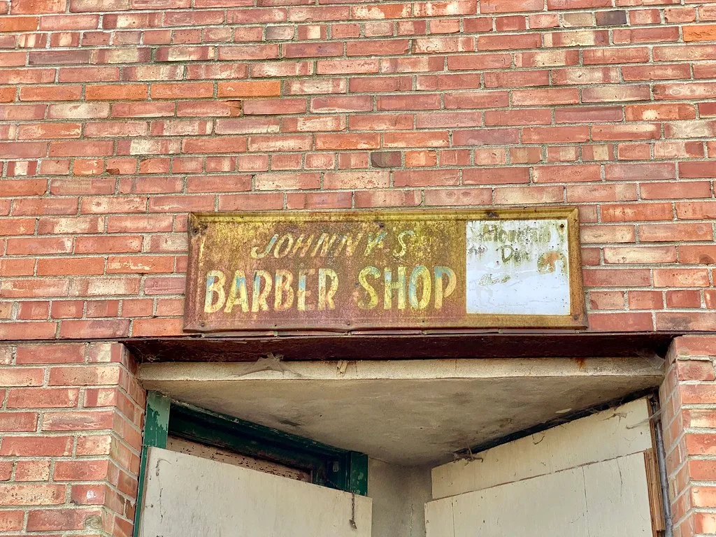 Entrée condamnée d’un ancien salon de coiffure sous une enseigne rouillée.