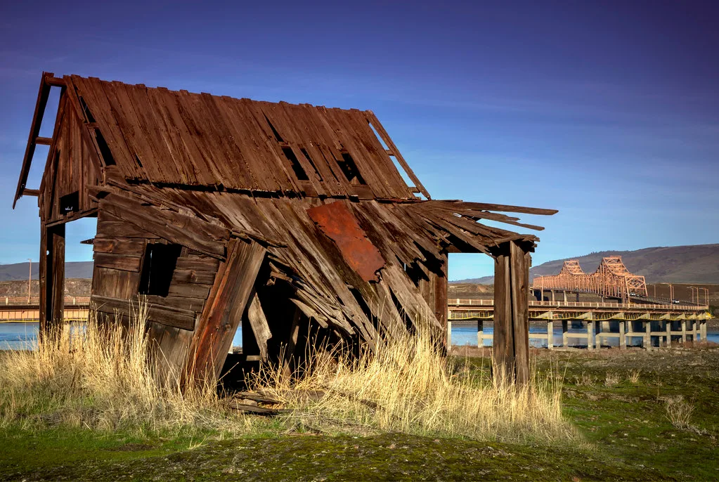 Ruined wooden hut near a bridge beside the water.
