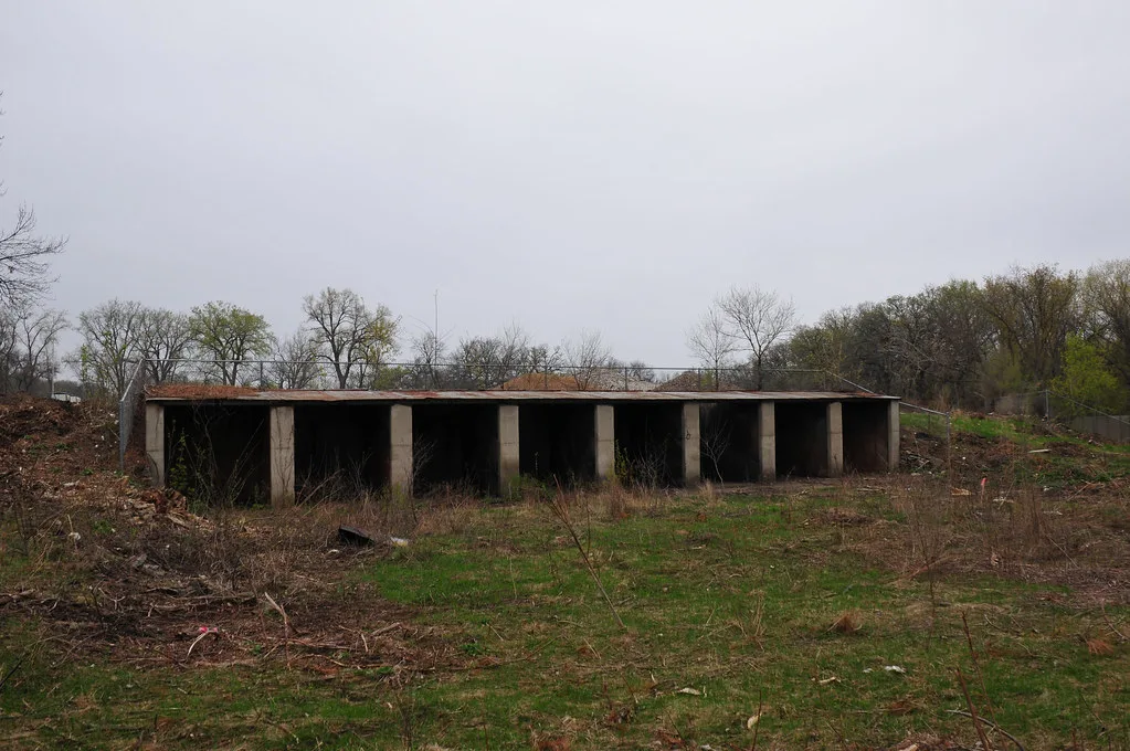Ancienne structure basse en béton aux travées ouvertes dans un terrain envahi d’herbes.