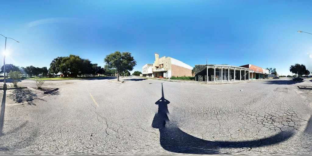 Ancien cinéma fermé et bâtiments bas devant un parking vide au sol fissuré.