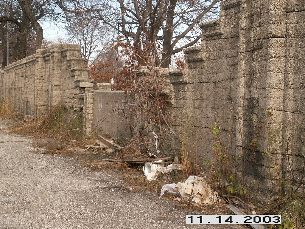 Abandoned grey block boundary wall, partially collapsed and overgrown with vines, with boards and litter at its foot.