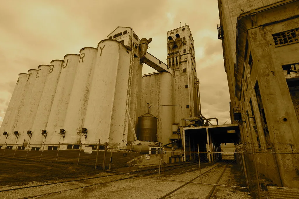 Large pale silos connected to deteriorated industrial towers with railway tracks in the foreground.