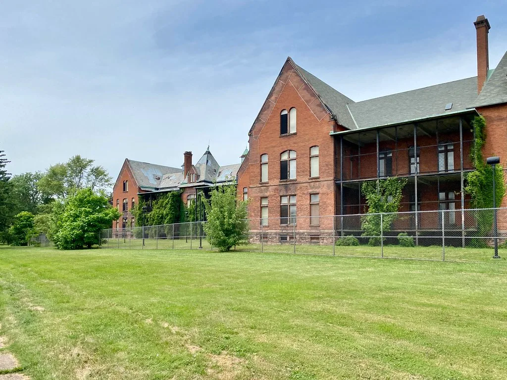 Old brick complex with boarded windows behind a fence.