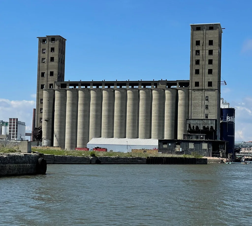 Abandoned concrete silos between two towers with empty windows beside the water.