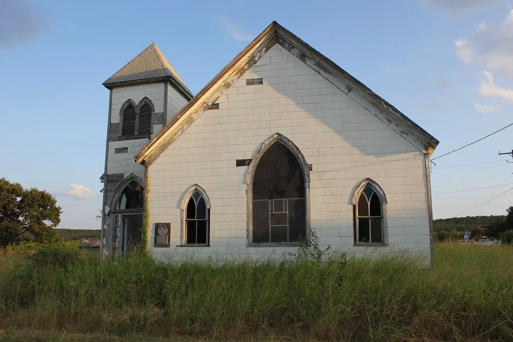 Old white church with broken windows in tall grass.
